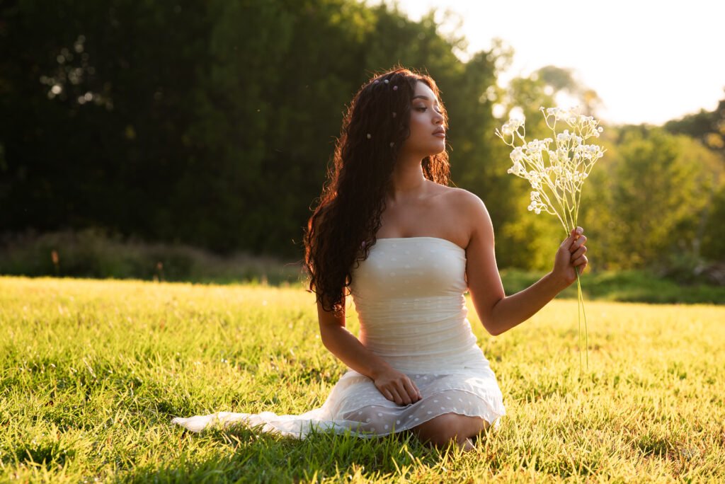 Portrait of a woman in a white strapless textured top, standing in a sunlit grassy field at golden hour. Her long, dark curly hair is dotted with tiny purple and white flowers and glows at the edges from the warm backlight. With her eyes lowered, she looks toward a bunch of white baby's breath held up in her left hand. The background is a soft, blurred wash of green trees and bright sky.