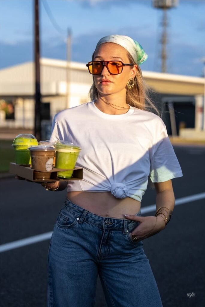 A model (woman) wearing a stylish top and Blue jeans with orange tinted sunglasses holding 3 matcha drinks in her hands